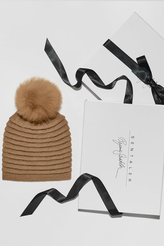 Sentaler Adult Ribbed Hat With Oversized Fur Pompon featured in Baby Alpaca and available in Dark Camel. Seen as off figure beside Sentaler box.